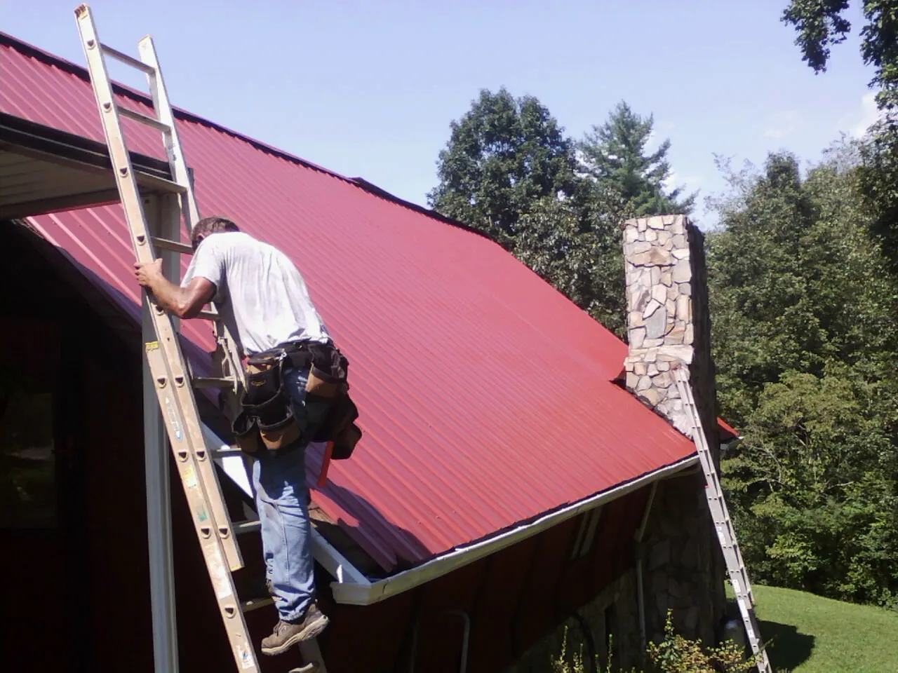 Roofer on a ladder beside a red metal roof and stone chimney — Soffit & Fascia Repair by Roofing Brothers in Wallingford